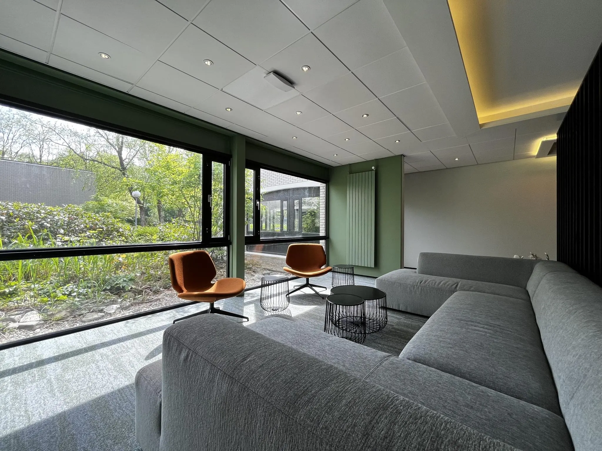 Moderne wachtruimte met grijze hoekbank, twee oranje stoelen en grote ramen met uitzicht op een groene tuin bij Verderlaan.